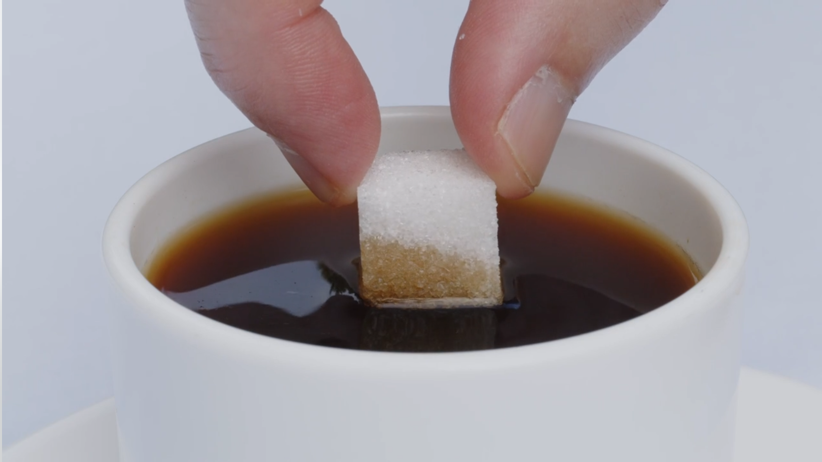 Zwei Finger halten ein Stück Würfelzucker in eine Tasse, die mit Kaffee gefüllt ist. Der Zuckerwürfel saugt den Kaffee entgegen der Schwerkraft von unten nach oben.